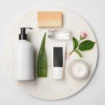 Top view of different cosmetic containers, soap, aloe vera leaf and rose flower on white round surface